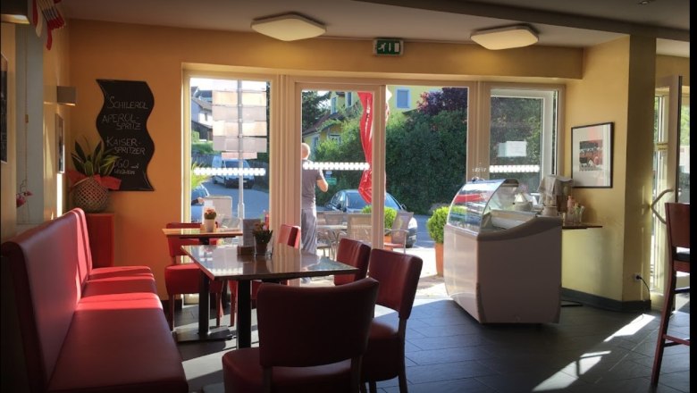 Innenbereich einer Bäckerei mit roten Sitzbänken und Tischen, großen Fenstern und einer Eistheke.