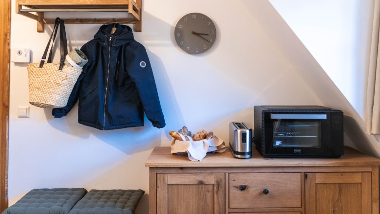 Ein Flur mit einer Holzgarderobe, einer Jacke, einem Hut, einer Tasche, einem Toaster, einem Ofen und einem Brotkorb auf einem Holzschrank.