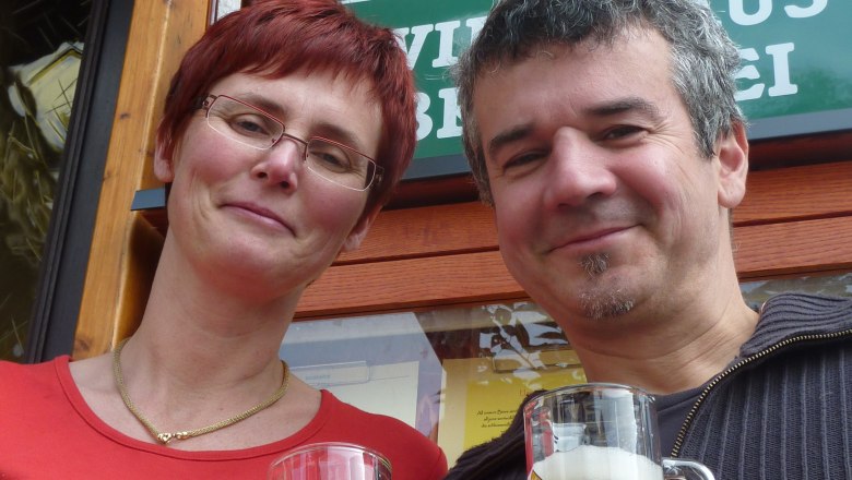 Zwei Personen stoßen mit Bierkrügen an vor einem Schild der Wirtshausbrauerei Haselböck.