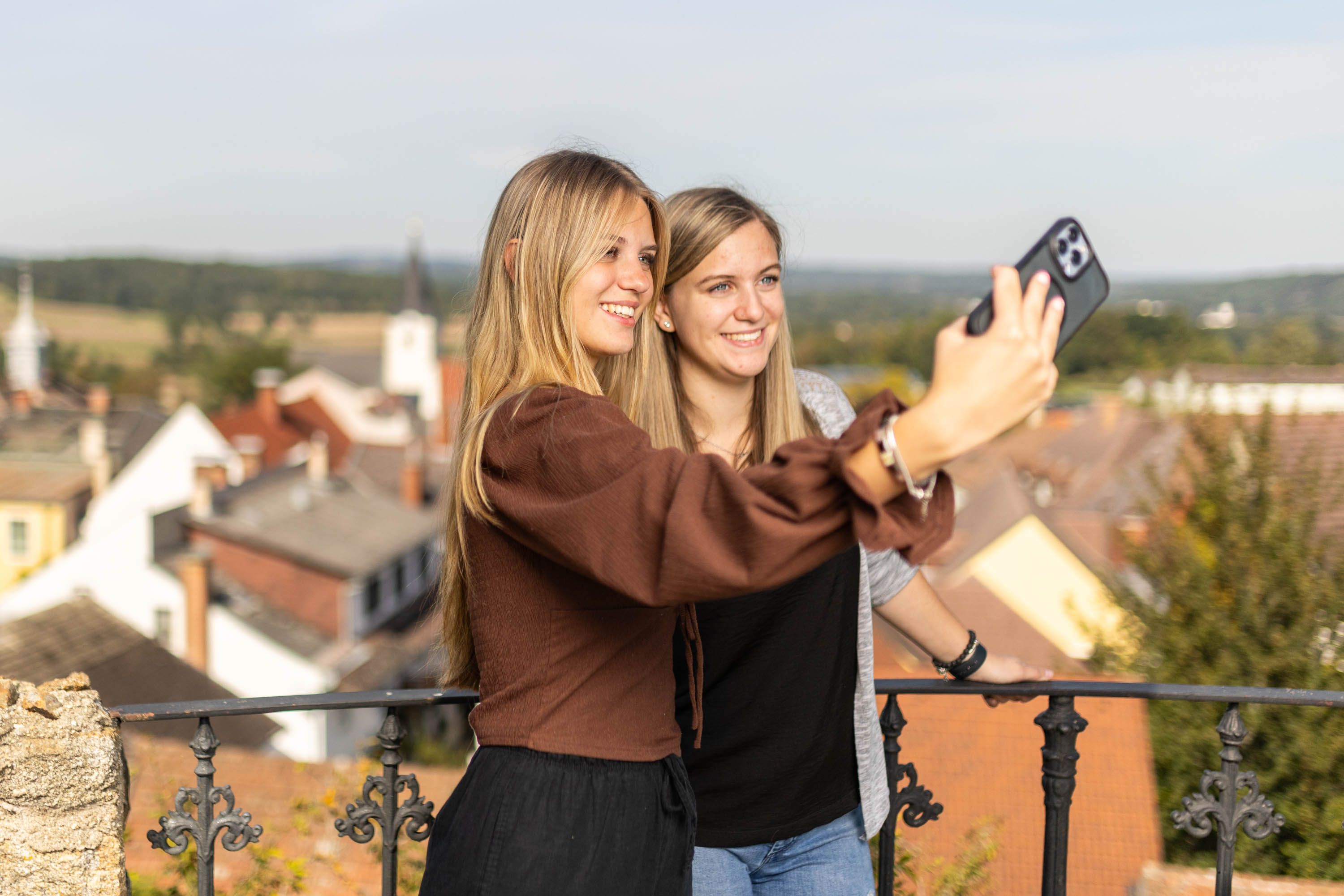 Zwei Frauen machen ein Selfie auf einem Balkon mit Blick auf eine Stadt.