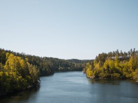 Die sanften Wellen des Stausees Ottenstein spiegeln die &uuml;ppige gr&uuml;ne Landschaft wider, w&auml;hrend die Sonne am klaren Himmel strahlt. Umgeben von dichten W&auml;ldern und sanften H&uuml;geln l&auml;dt dieser Ort dazu ein, die Ruhe der Natur zu genie&szlig;en und die frische Luft beim Radfahren zu erleben.