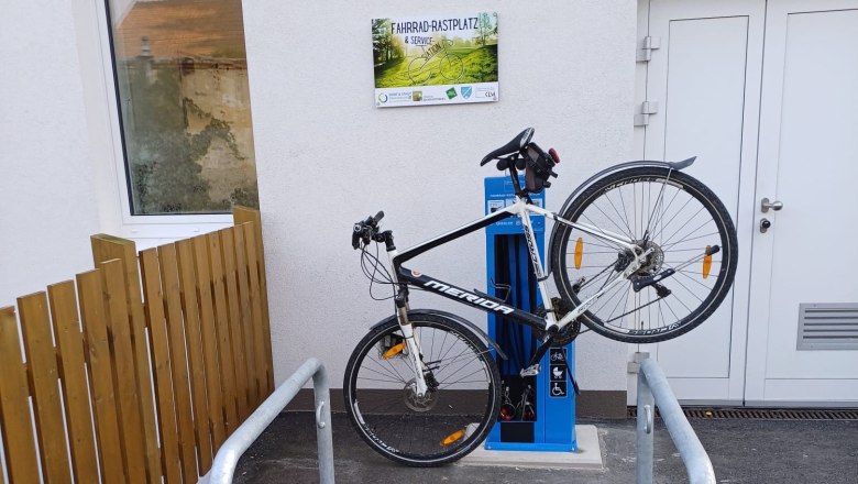 Fahrrad an einer blauen Service-Station befestigt, neben einem Geb&auml;ude mit einem Schild 'Fahrrad-Rastplatz & Service Station'.