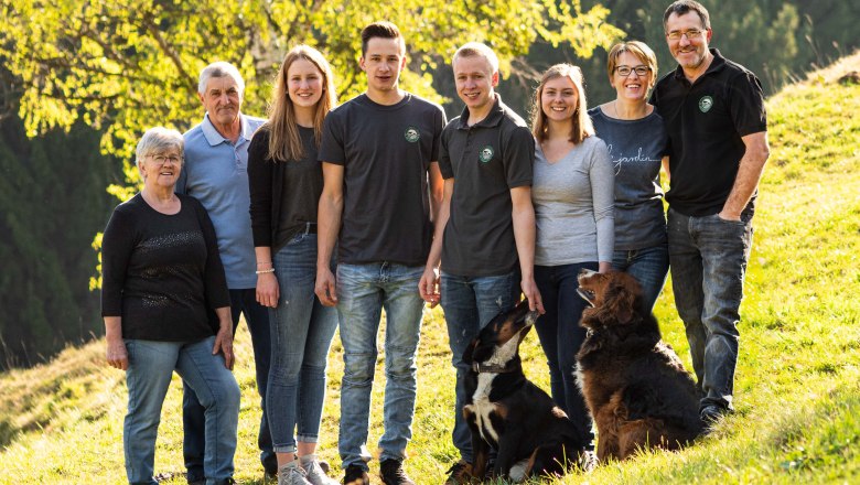 Eine gro&szlig;e Familie mit zwei Hunden steht auf einer gr&uuml;nen Wiese vor einem Baum.