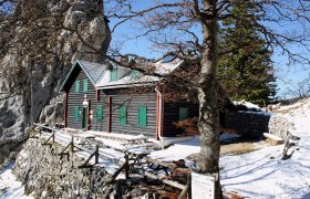 Eine Berghütte aus Holz mit grünen Fensterläden, umgeben von Schnee und Felsen, unter einem klaren blauen Himmel.