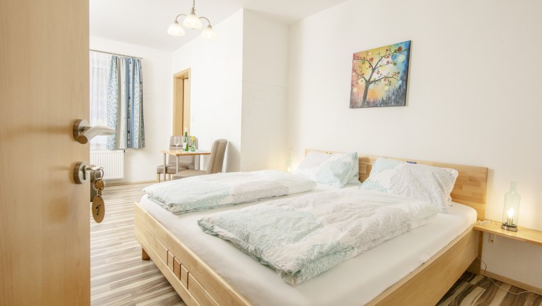 Ein helles Gästezimmer mit Doppelbett, Tisch, Stühlen und einem bunten Wandbild.