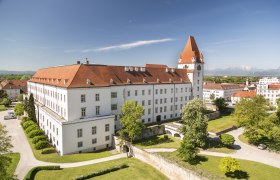 Blick auf eine historische Militärakademie mit rotem Dach und Turm, umgeben von grünen Bäumen und Rasenflächen, im Hintergrund Berge.