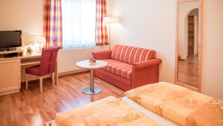 Gem&uuml;tliches G&auml;stezimmer mit Doppelbett, rotem Sofa, Schreibtisch, Fernseher und gro&szlig;em Spiegel.