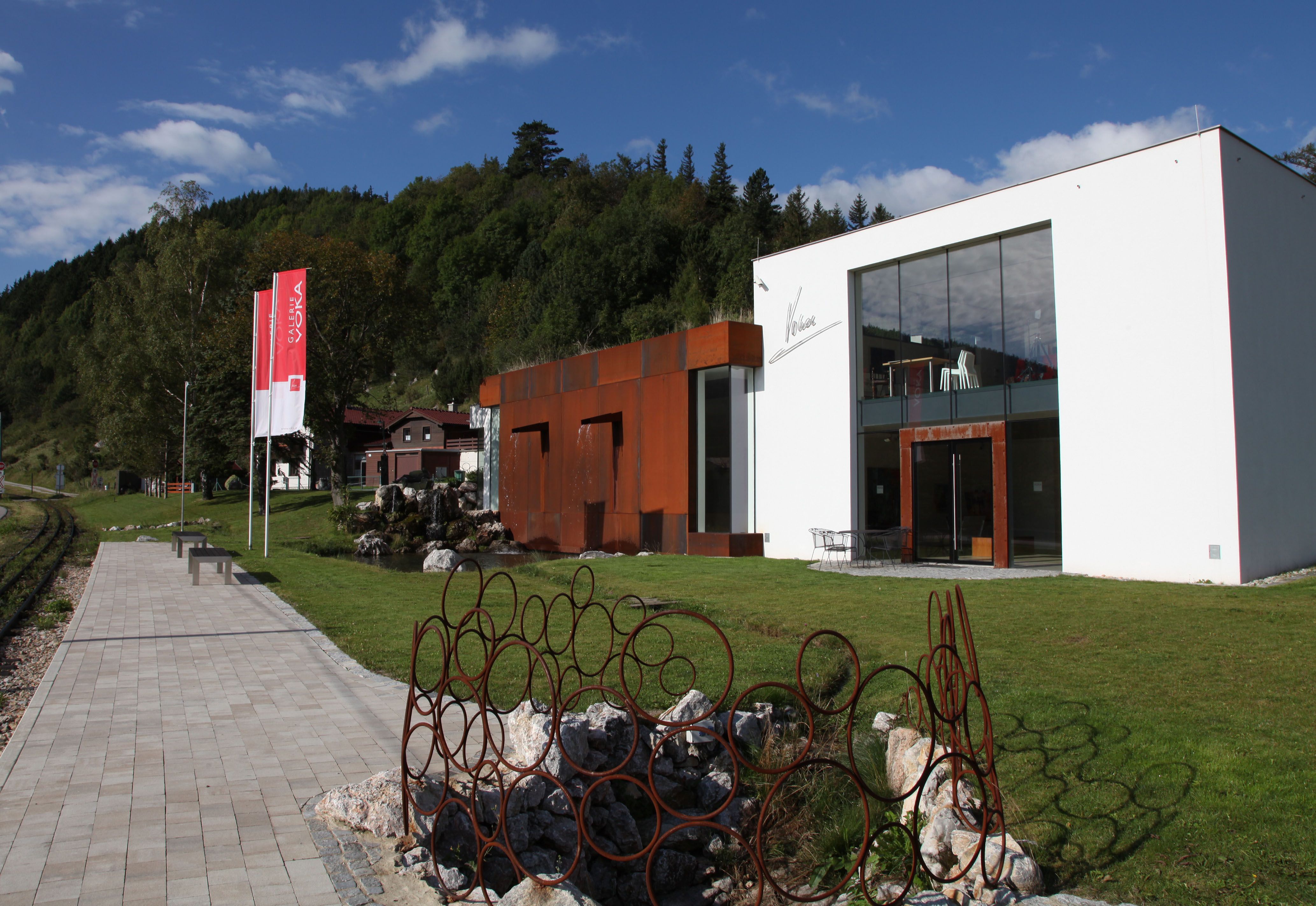 Moderne Galerie mit Glasfassade und rostigen Metallskulpturen im Grünen.