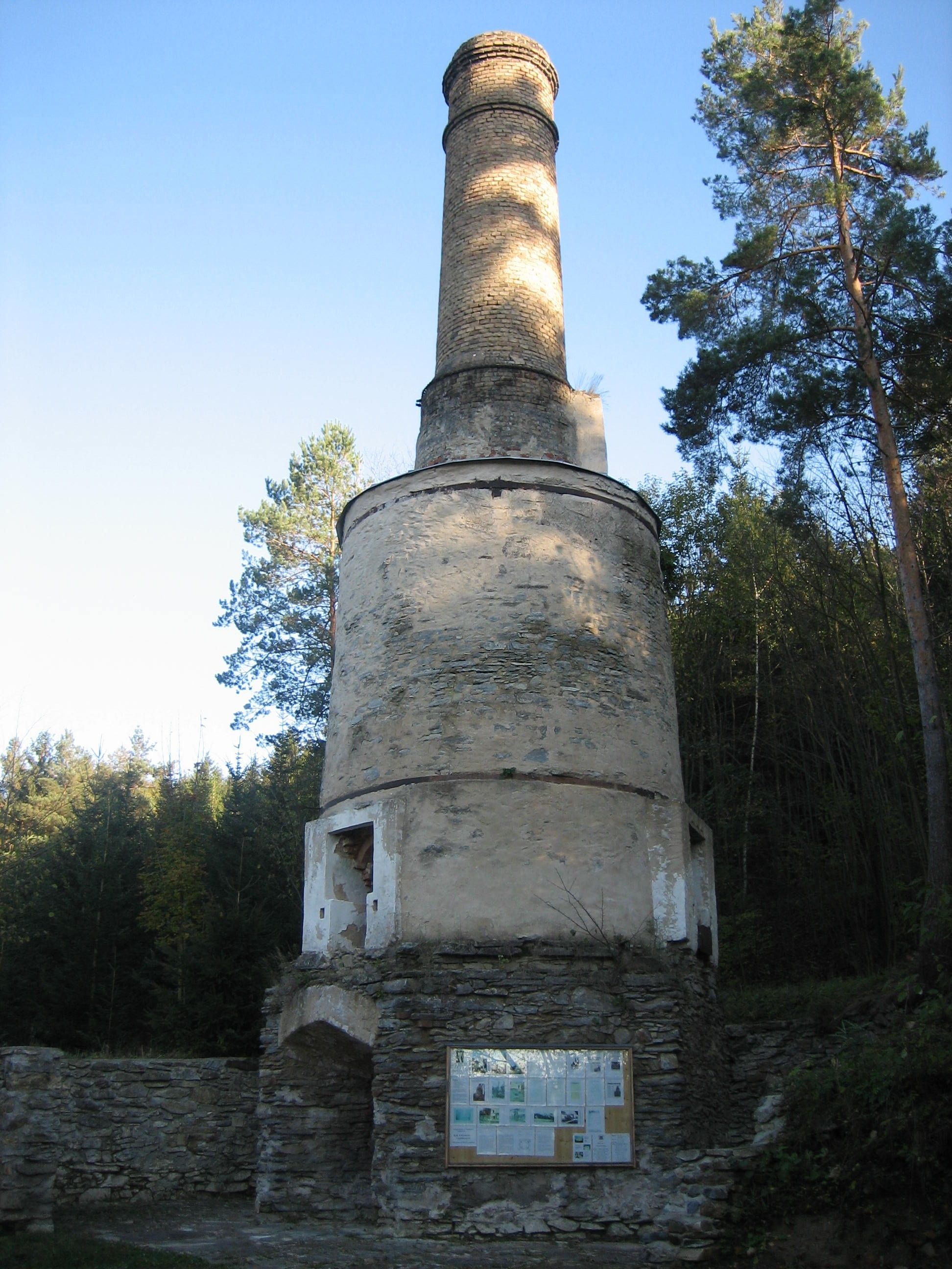 Ein alter, runder Schornstein aus Stein in einem Waldgebiet mit Informationstafel.