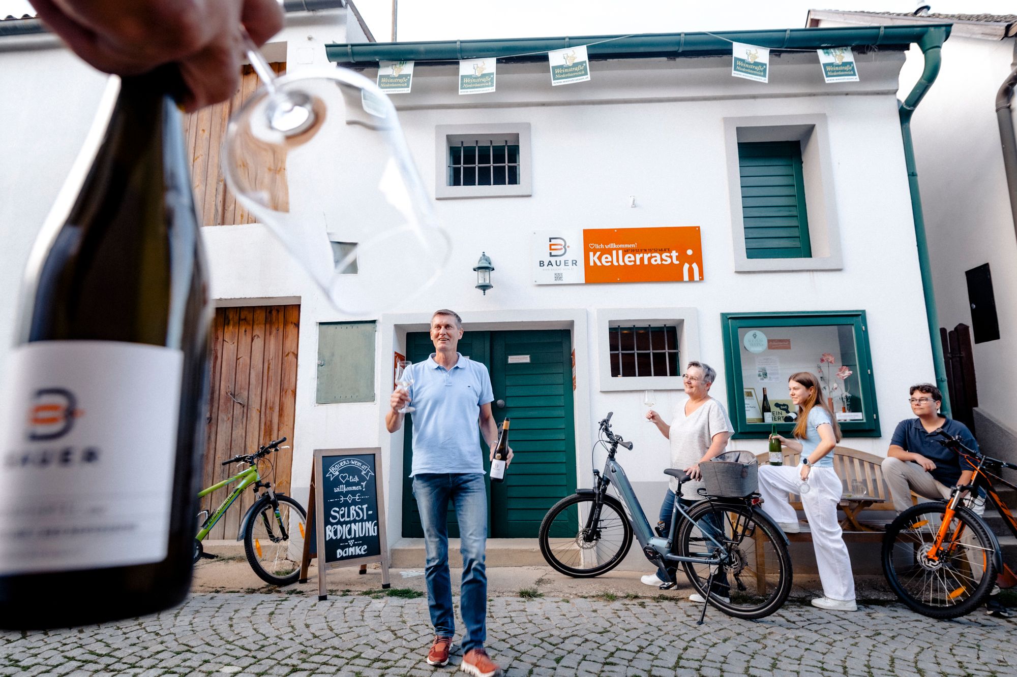 Menschen stehen vor einem Gebäude mit einem Schild 'Kellerrast', während eine Person im Vordergrund eine Weinflasche und ein Glas hält.