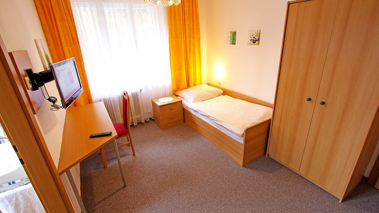 Ein einfach eingerichtetes Einzelzimmer mit Bett, Schreibtisch, Stuhl, Kleiderschrank und Fernseher. Gelbe Vorh&auml;nge und Teppichboden.