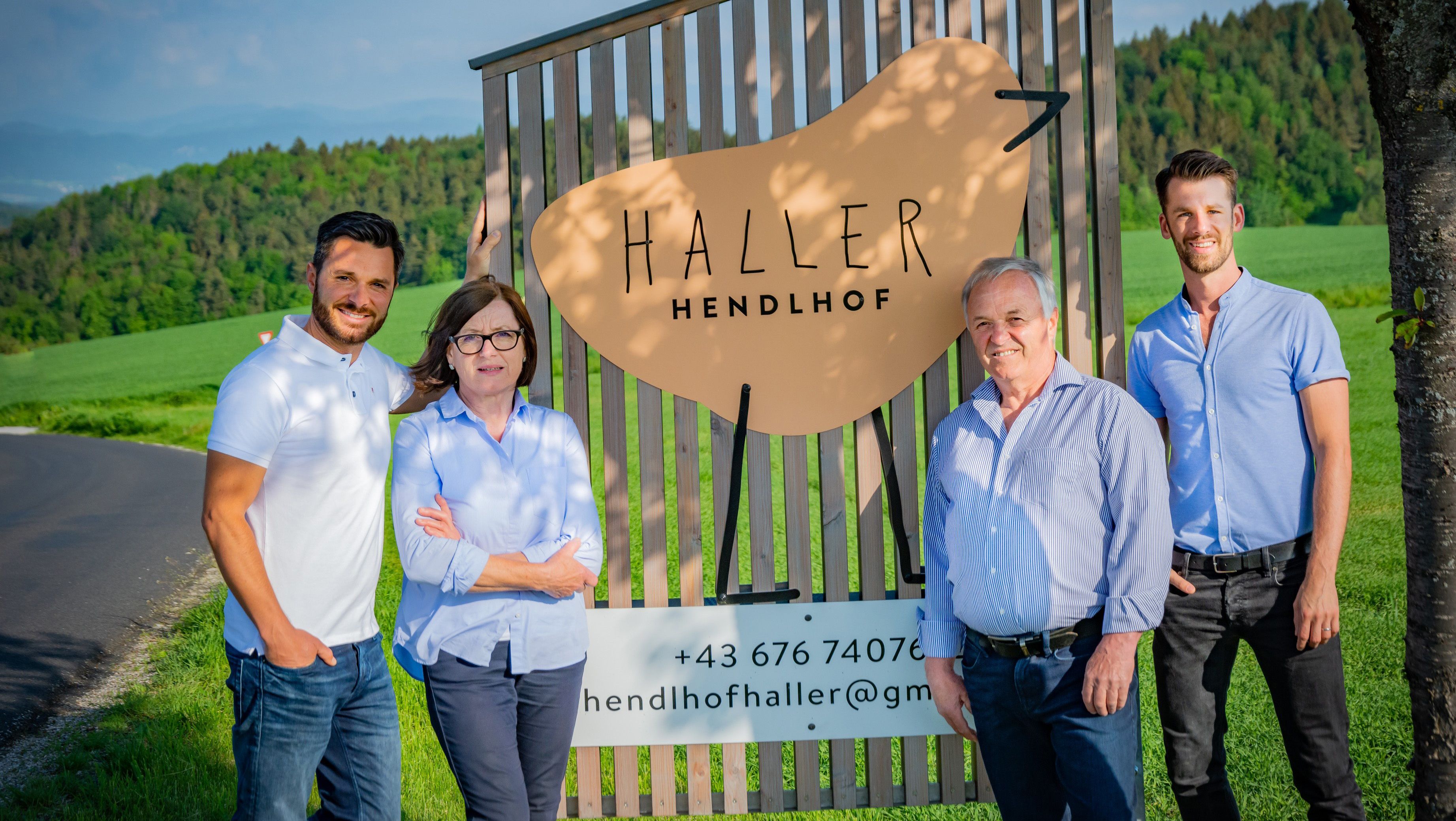VIer Personen stehen vor einem Holzschild mit einem Logo vom Haller Hendlhof in einer ländlichen Umgebung.