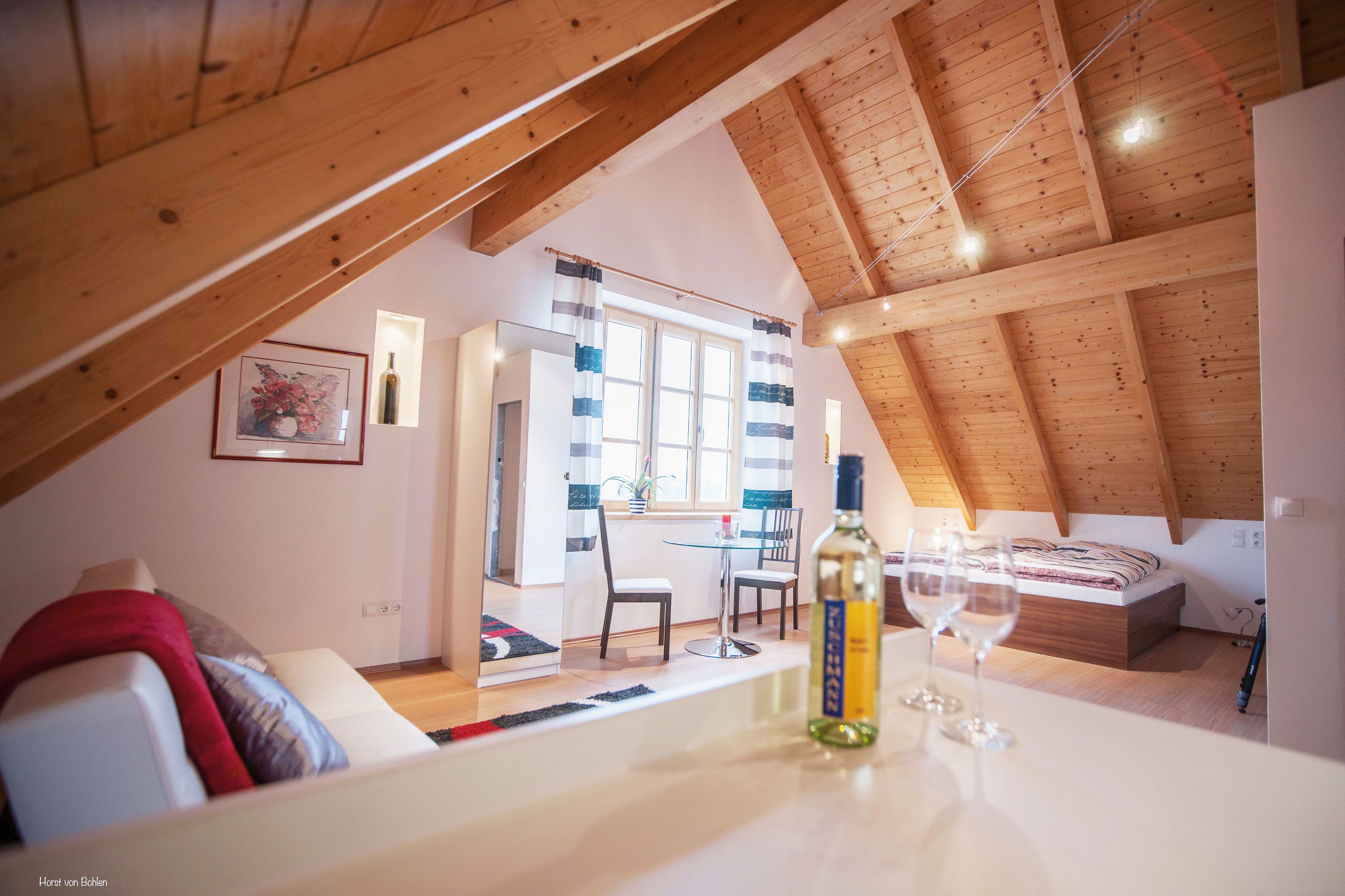 Gemütliches Dachgeschosszimmer mit Holzbalkendecke, Bett, Sofa, Tisch und Weinflasche.