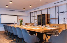 Moderner Konferenzraum mit gro&szlig;em Holztisch, grauen St&uuml;hlen und Pr&auml;sentationsleinwand.