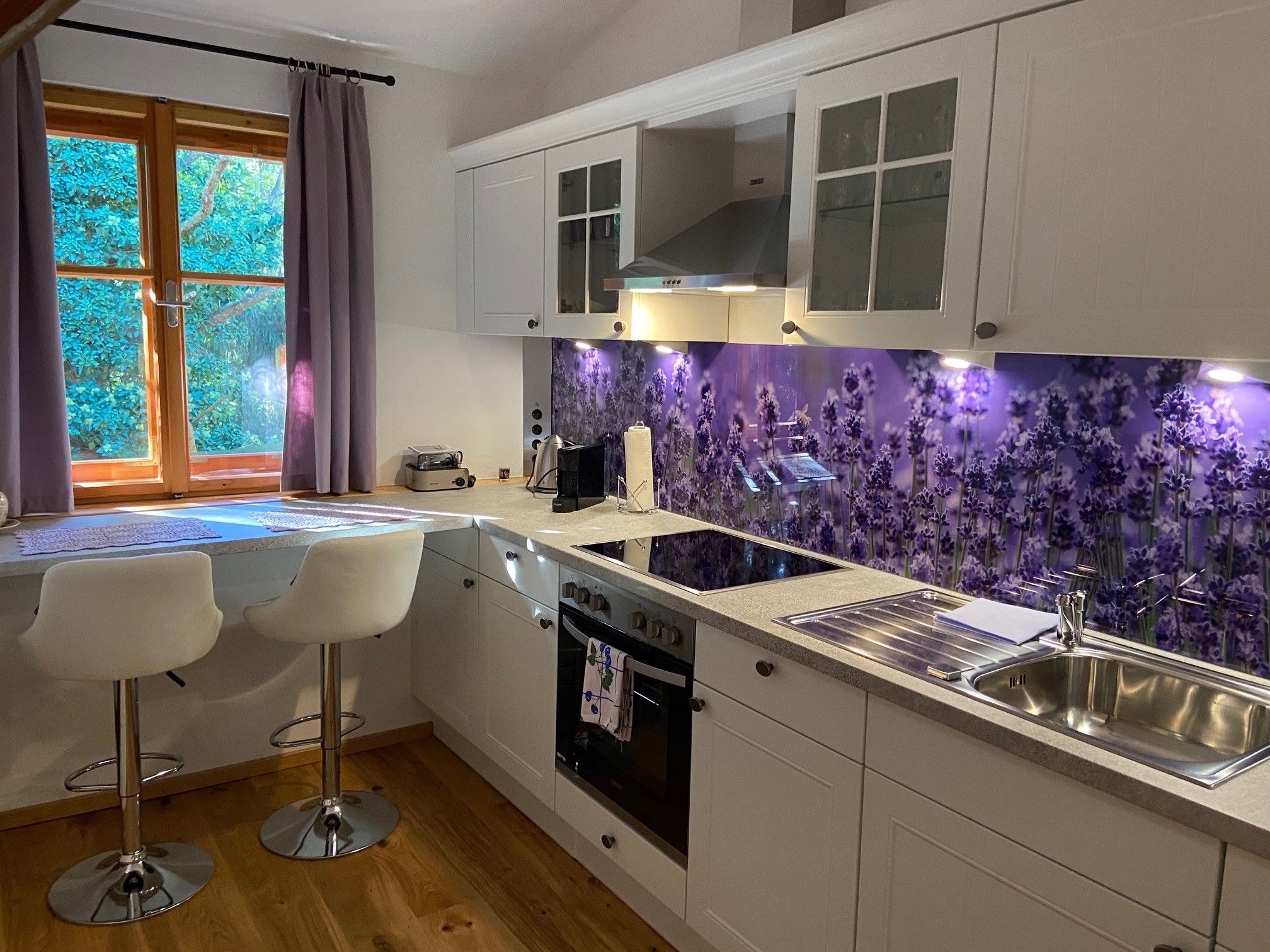 Moderne Küche mit weißen Schränken, Lavendelrückwand und zwei Barhockern vor einem Fenster mit lila Vorhängen.