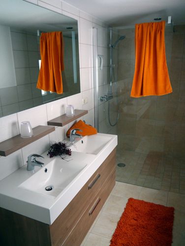 Modernes Badezimmer mit Doppelwaschbecken, großer Spiegel, Dusche und orangefarbenen Akzenten.
