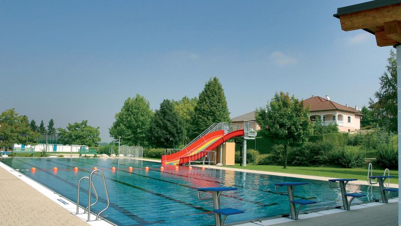 Ein Freibad mit einem Schwimmbecken, einer roten Rutsche und Sprungbrettern. Im Hintergrund sind B&auml;ume und ein Haus zu sehen.