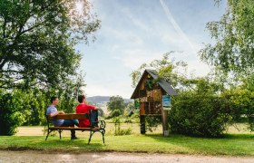 Zwei Personen sitzen auf einer Bank im Gr&uuml;nen vor einem Insektenhotel.