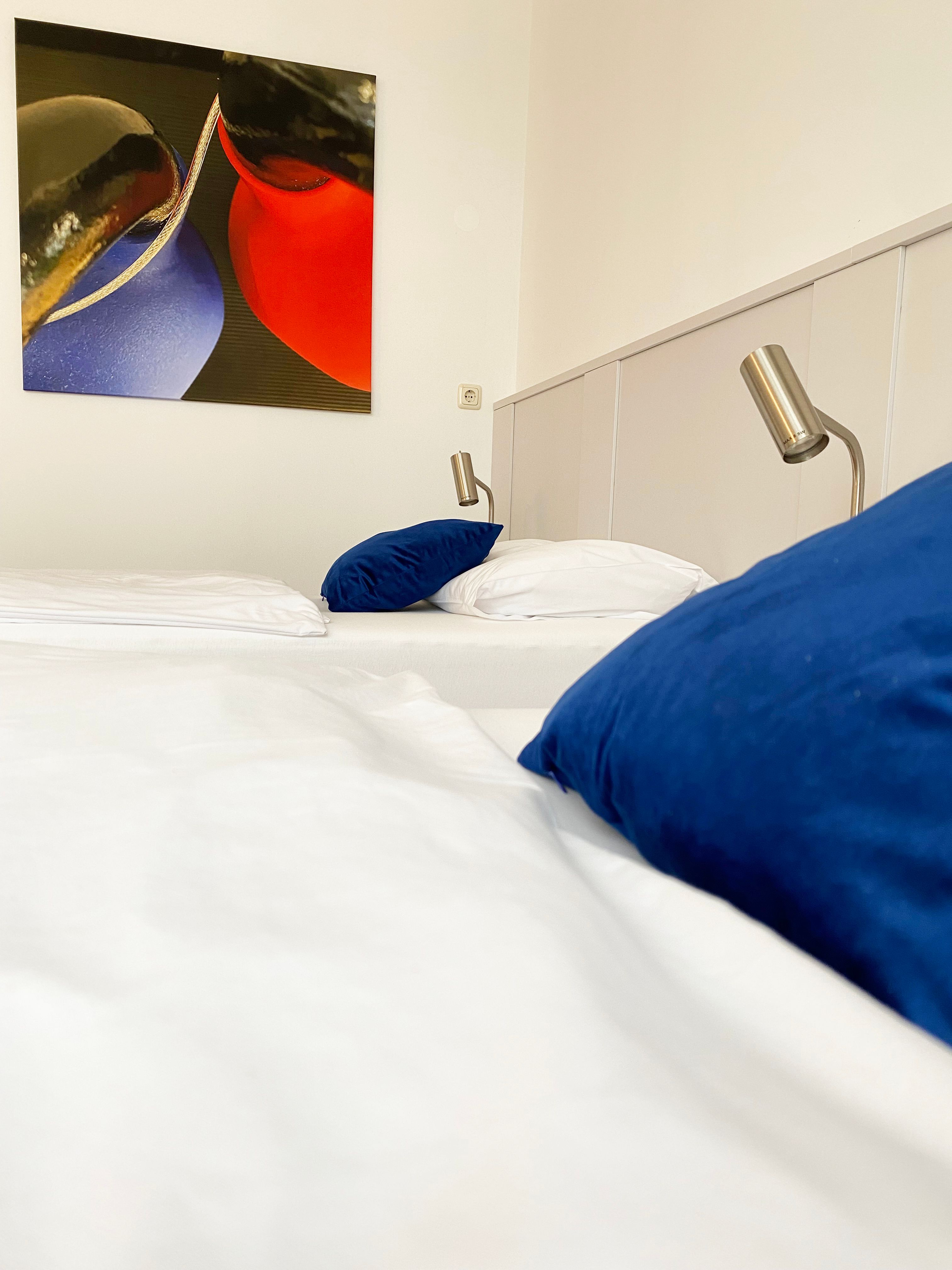 Ein Gästezimmer mit einem Bett, blauen Kissen, Leselampen und einem abstrakten Gemälde an der Wand.