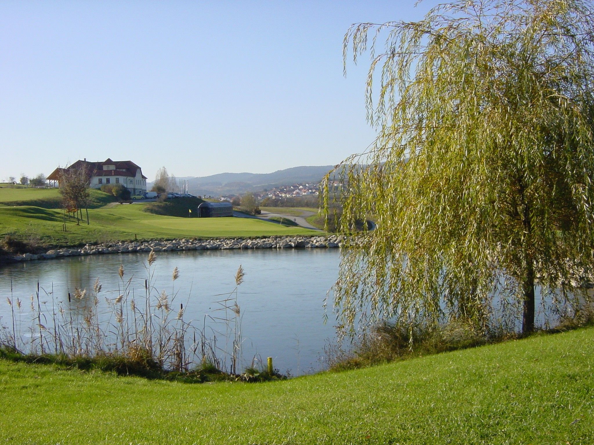 Golfplatz mit Teich, Bäumen und einem Gebäude im Hintergrund.