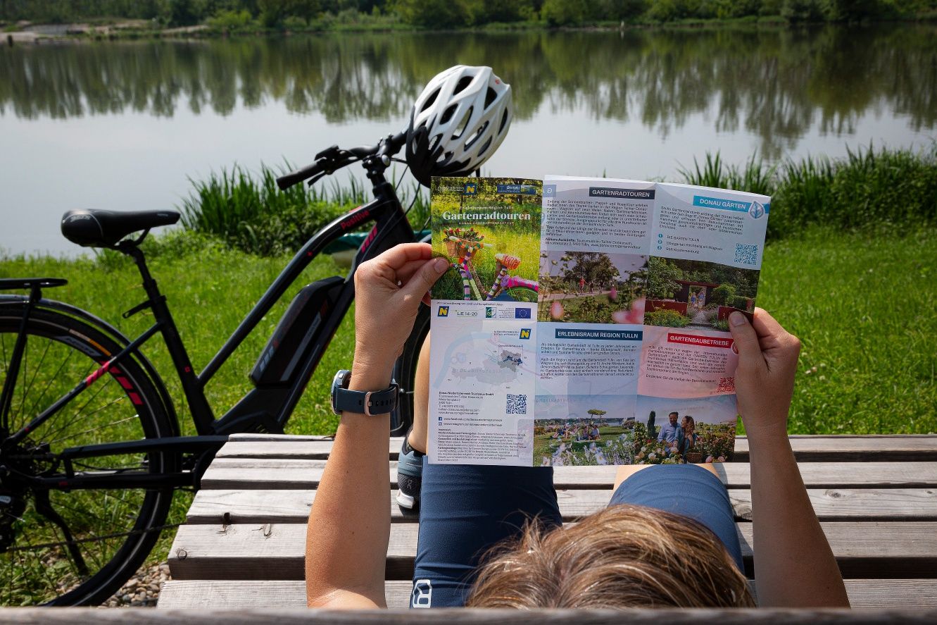 Person liest eine Faltkarte über Gartenradtouren neben einem Fahrrad am See.