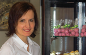 Eine Frau mit weißer Bluse steht lächend neben einer Vitrine mit verpackter Mehlspeise.