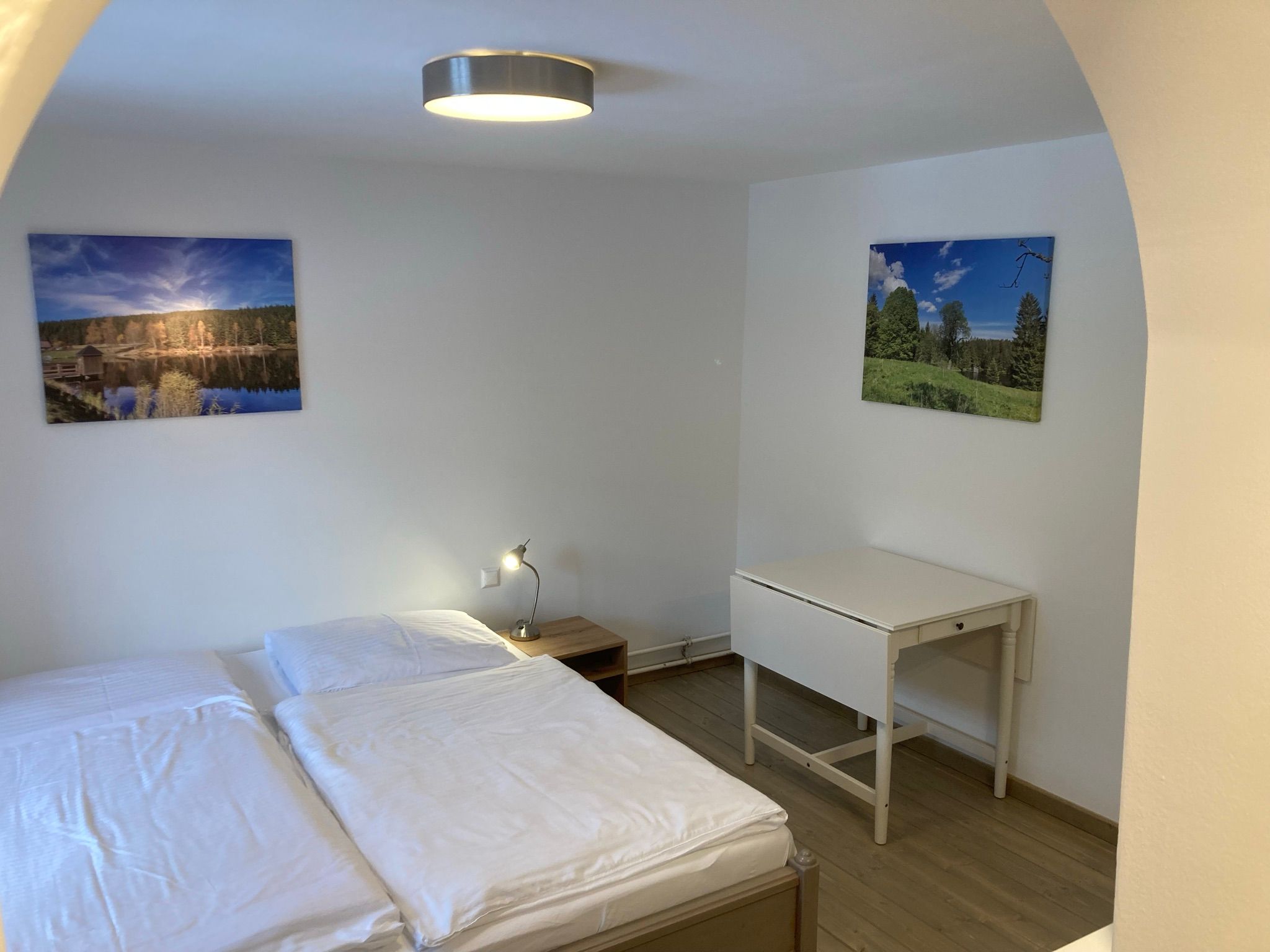 Ein schlicht eingerichtetes Zimmer mit einem Doppelbett, einem kleinen Tisch und zwei Landschaftsbildern an der Wand.