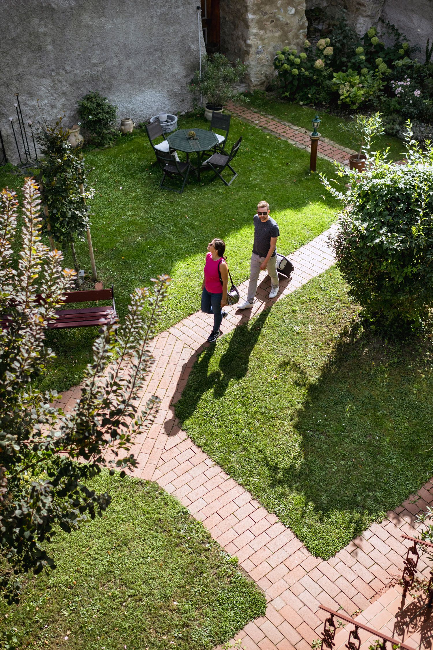 Zwei Personen gehen auf einem gepflasterten Weg durch einen grünen Garten mit Tisch und Stühlen.