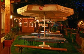 Ein gem&uuml;tlicher Biergarten bei Nacht mit Holztischen, blau karierten Tischdecken und gro&szlig;en Sonnenschirmen der Marke Stiegl.