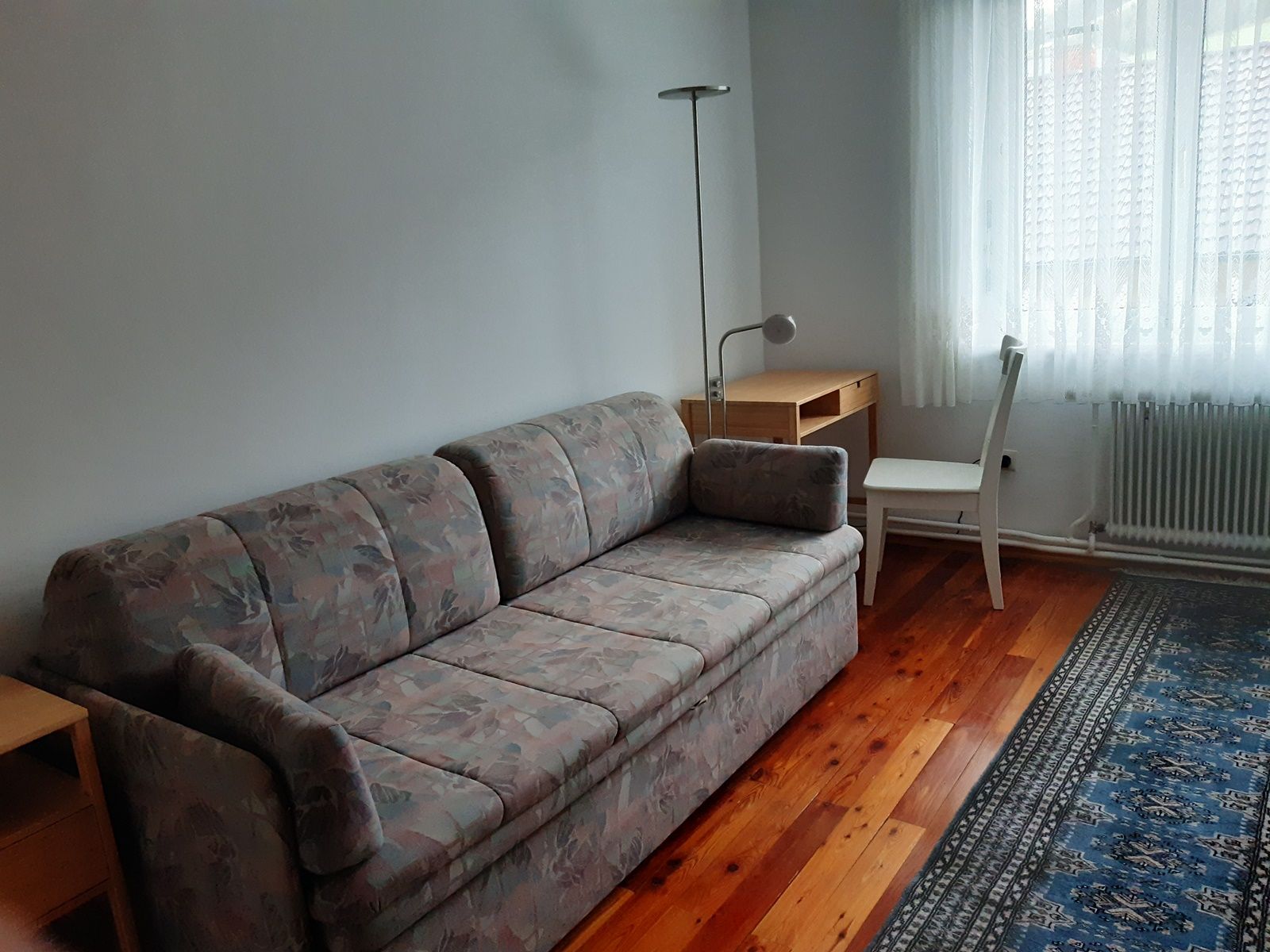 Ein schlicht eingerichtetes Zimmer mit einem Sofa, einem Schreibtisch, einem Stuhl und einem Teppich auf Holzboden.