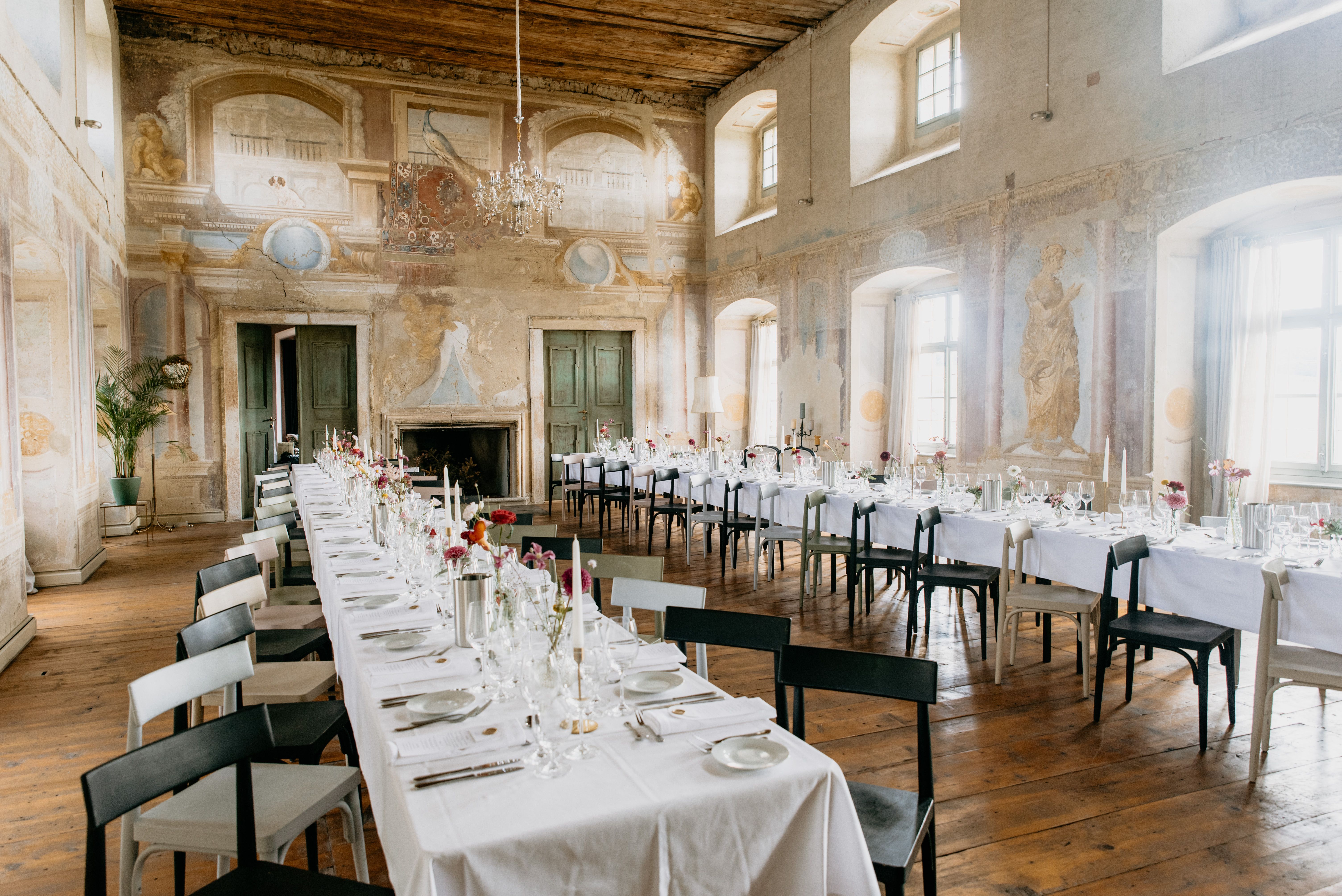 Eleganter Festsaal mit langen, gedeckten Tischen, antiken Wandmalereien und Kronleuchter.