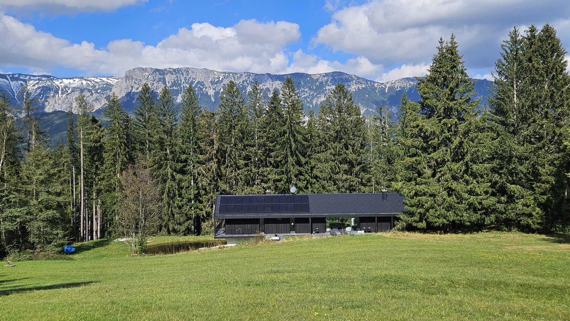 Modernes Haus mit Solarpanelen vor einem Wald und Bergen im Hintergrund.