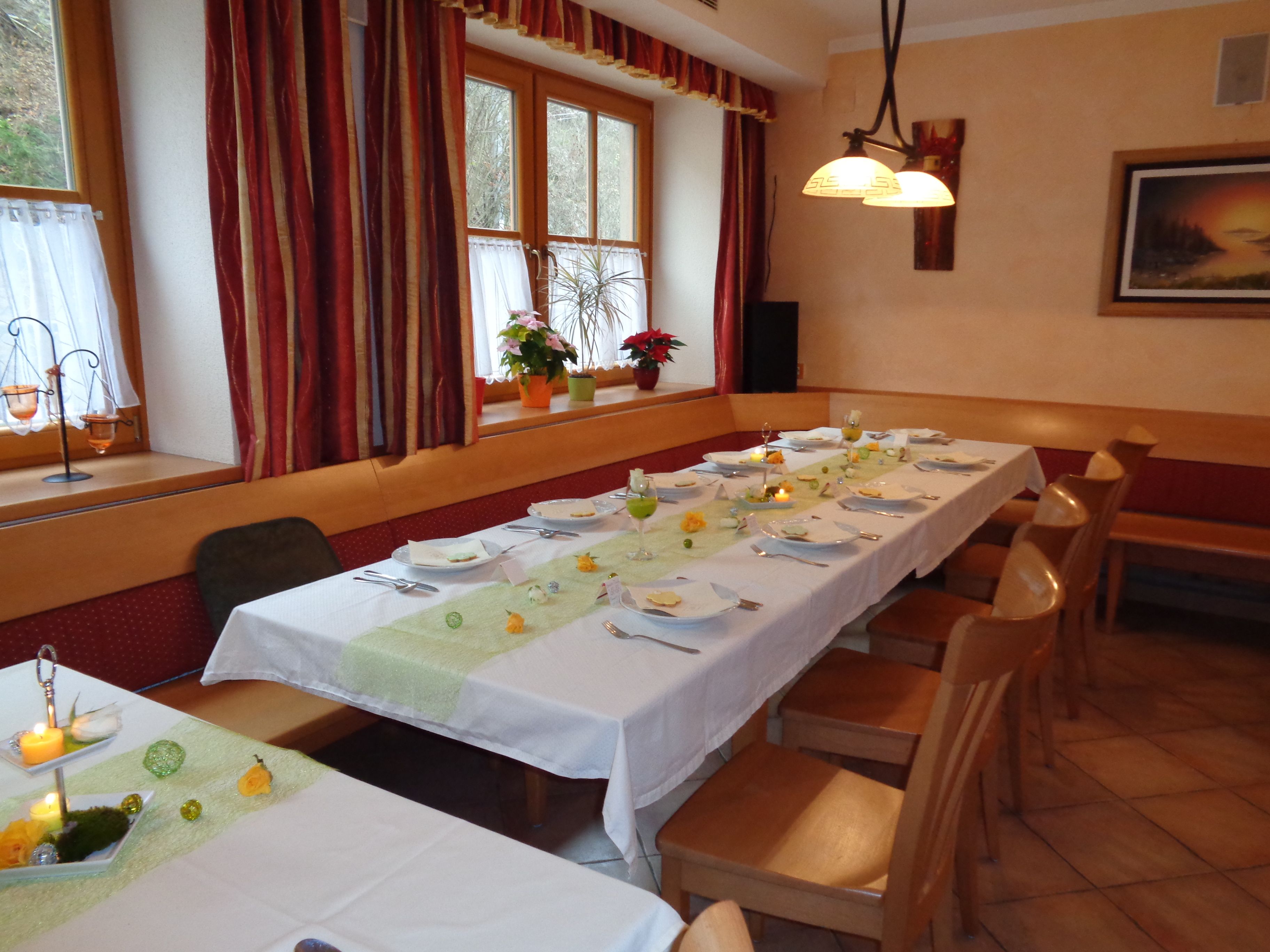 Ein festlich gedeckter Tisch in einem Gasthaus mit weißen Tischdecken, Kerzen und Blumen. Holzmöbel und Fenster mit Vorhängen im Hintergrund.