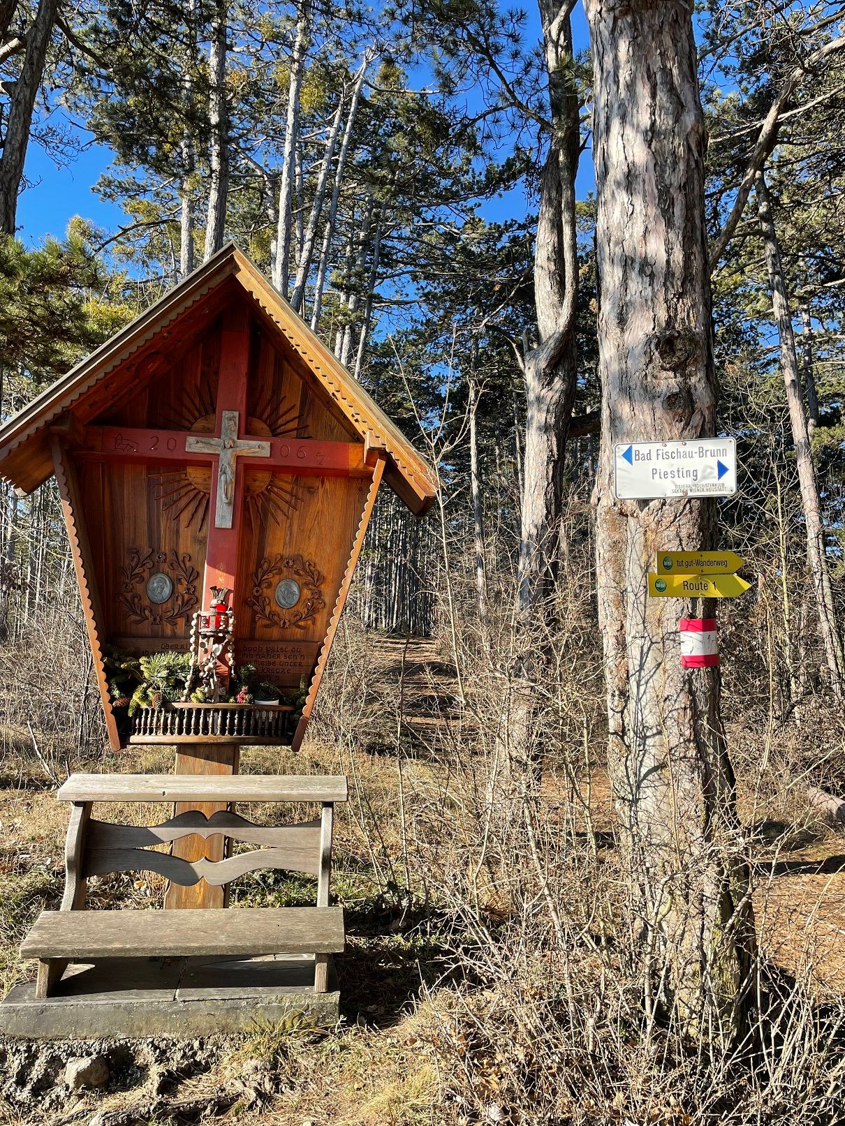 Holzkreuz mit Bank im Wald, daneben Wegweiser mit Richtungshinweisen.