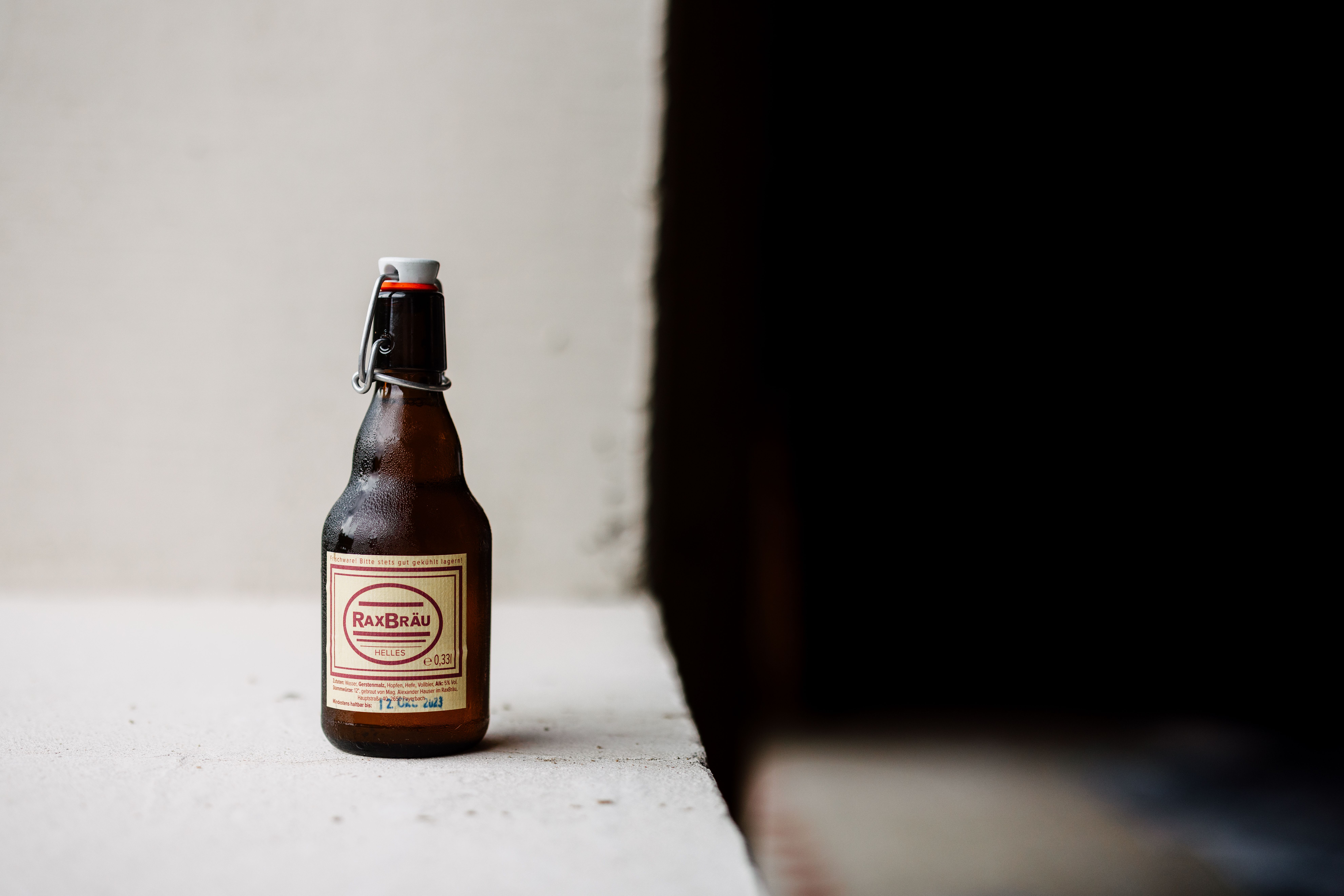 Eine Flasche RaxBräu Helles Bier auf einer weißen Oberfläche vor einem dunklen Hintergrund.
