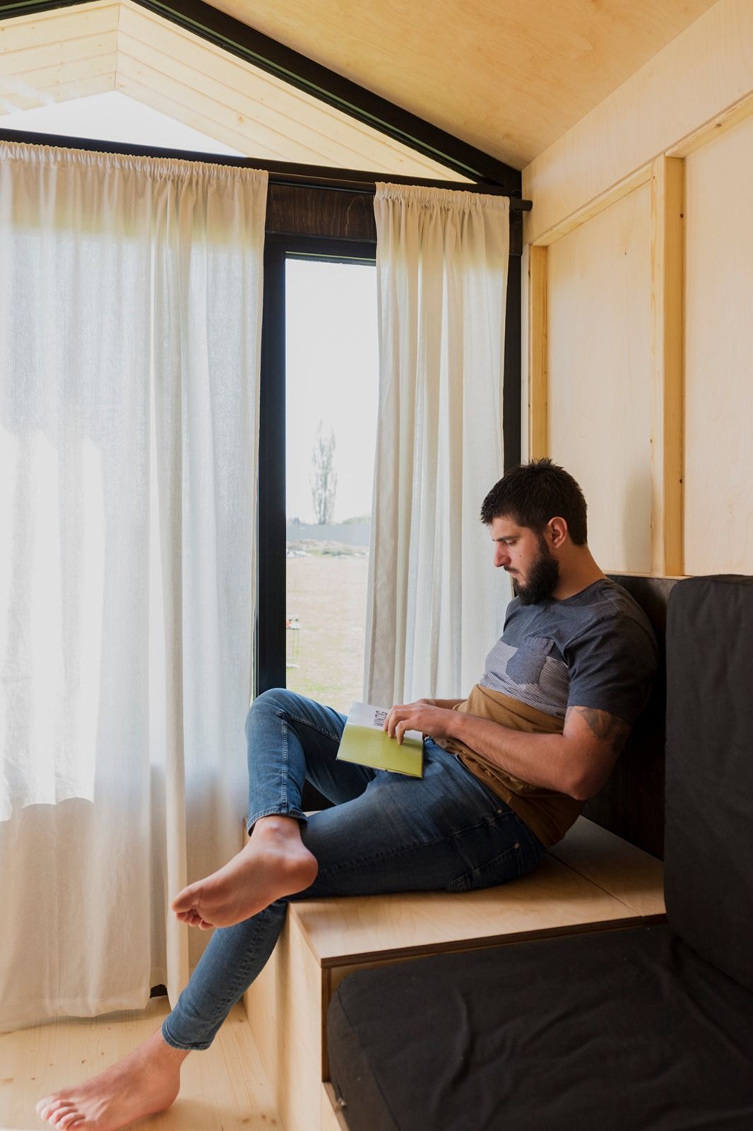 Ein Mann sitzt in einer gemütlichen Ecke einer Holzhütte und liest ein Buch. Durch das Fenster fällt Tageslicht herein.