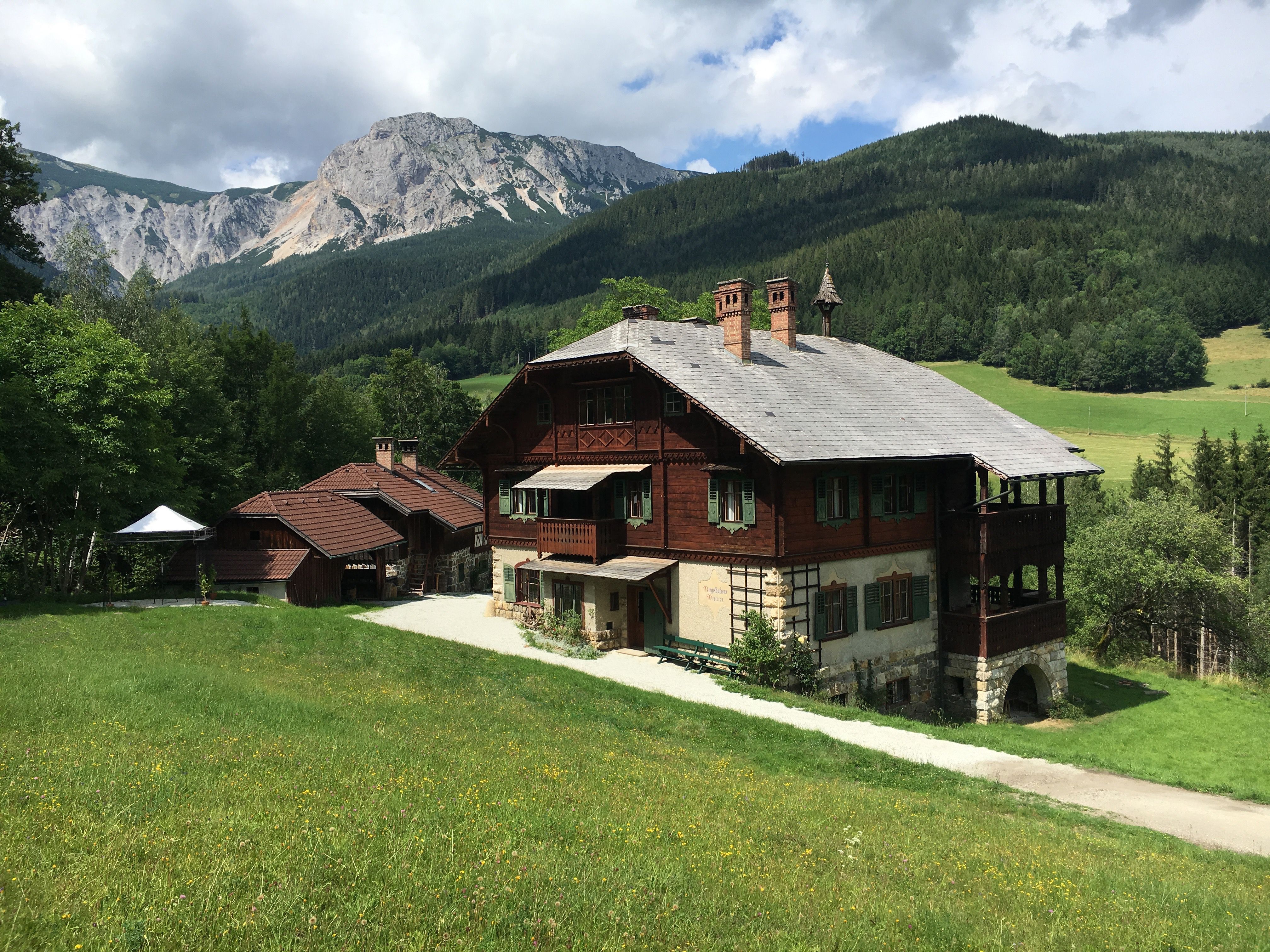 Ein traditionelles Holzhaus in einer bergigen Landschaft mit grünen Wiesen und bewaldeten Hügeln im Hintergrund.