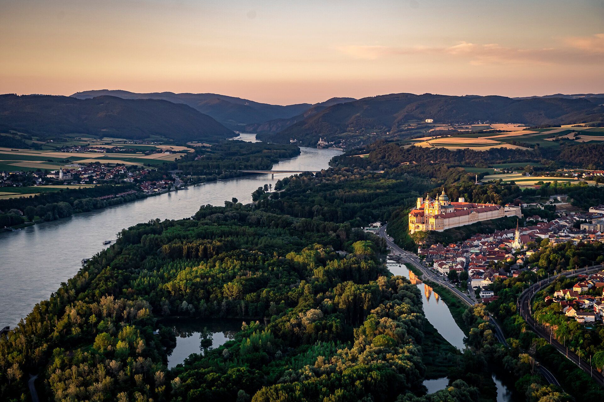 Die sanften Hügel und die glitzernde Donau umarmen das majestätische Stift Melk, während die goldenen Strahlen der Abendsonne die Landschaft in ein warmes Licht tauchen. Hier, wo Geschichte und Natur harmonisch verschmelzen, lädt die Umgebung zu unvergesslichen Erlebnissen ein.