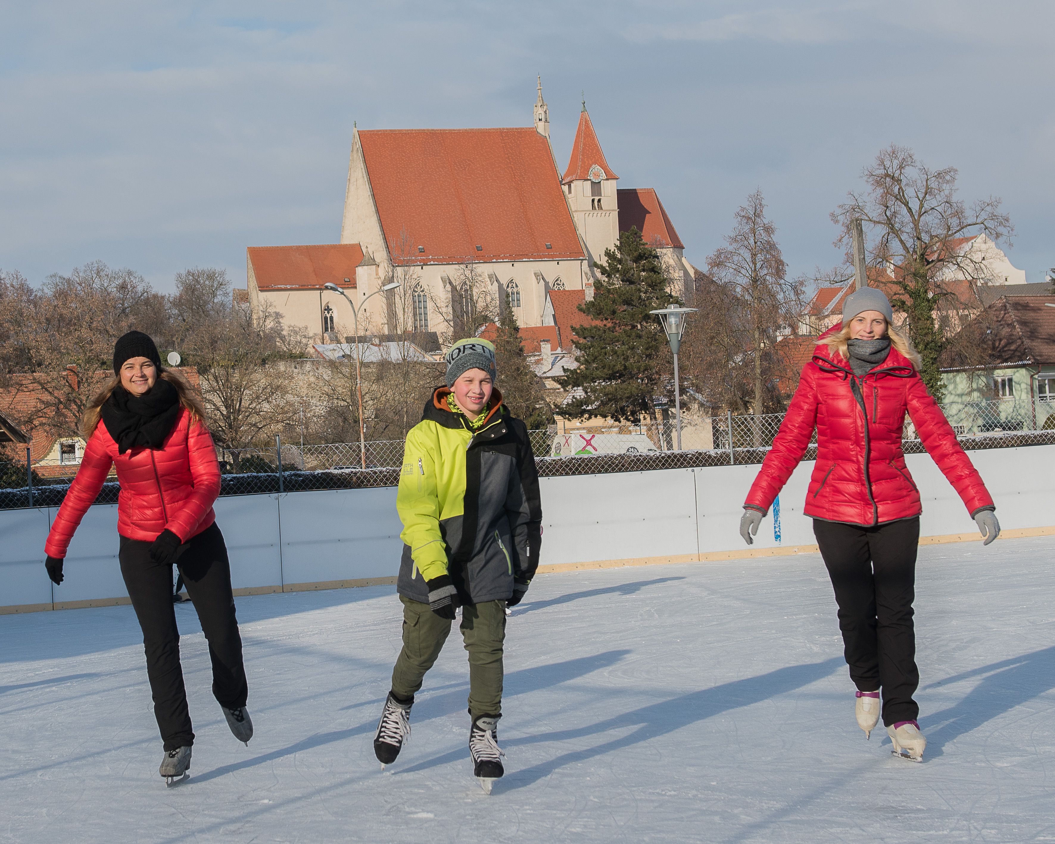 Drei Personen beim Eislaufen auf einer Eisbahn mit einer Kirche im Hintergrund.