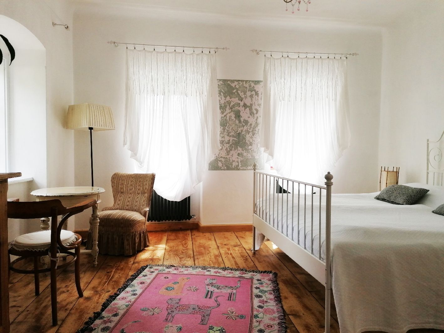 Helles Zimmer mit Holzboden, weißem Bett, zwei Fenstern mit Vorhängen, einem Tisch mit Stuhl und einem rosa Teppich.