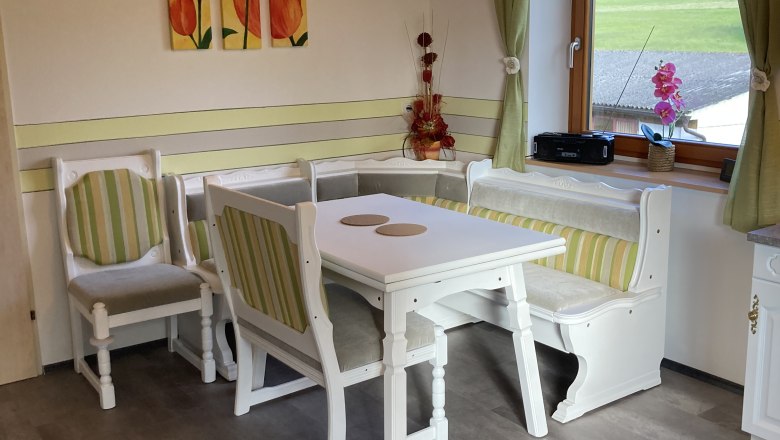 Gem&uuml;tliche Essecke mit Holzm&ouml;beln und gr&uuml;n-wei&szlig; gestreiften Polstern, Fenster mit Blick ins Gr&uuml;ne, Tulpenbild an der Wand.