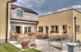 Ein gemütlicher Außenbereich mit Holztischen und -stühlen vor einem gelben Gebäude.