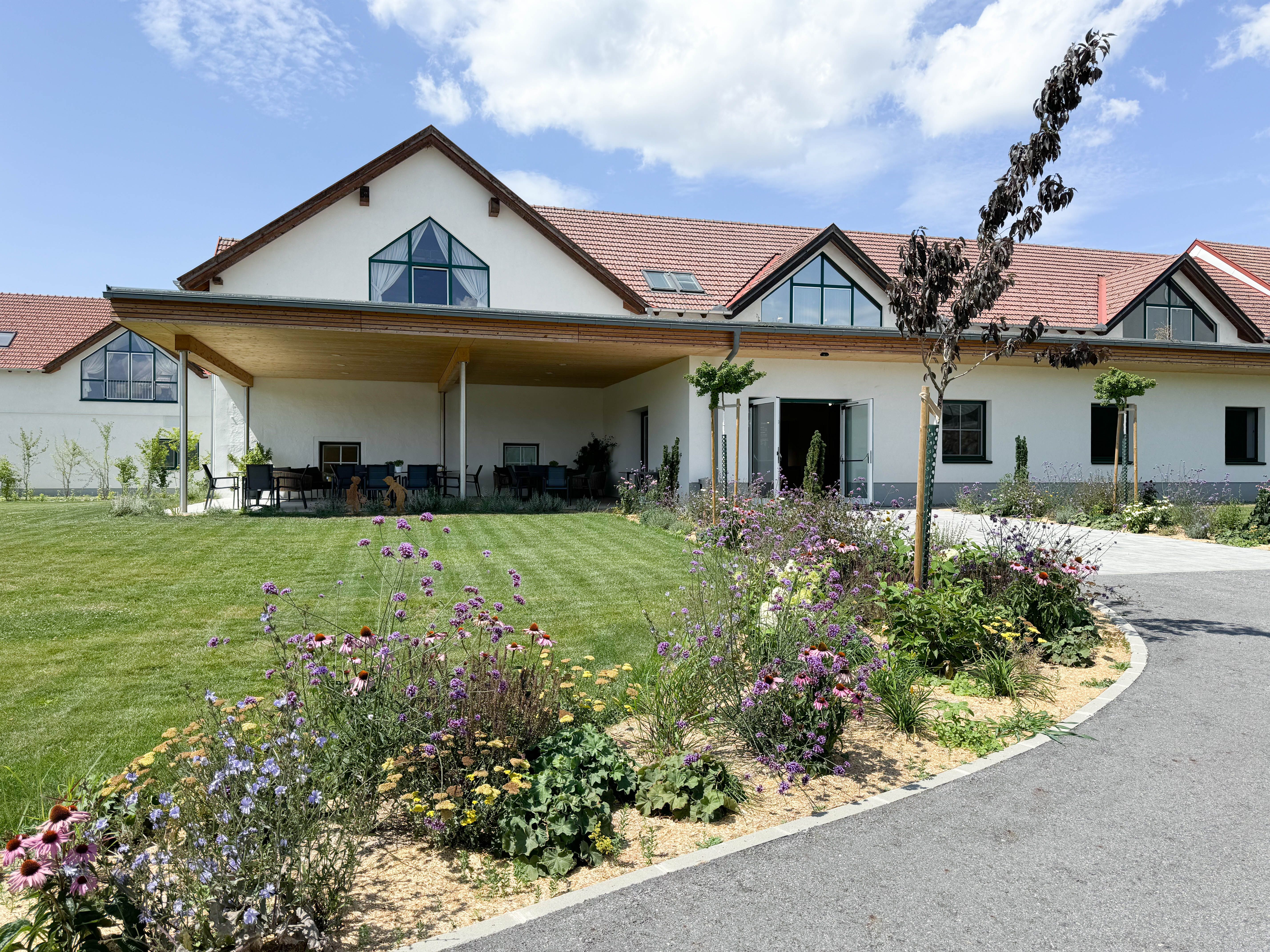 Ein modernes Gebäude mit rotem Dach, umgeben von einem gepflegten Garten mit bunten Blumen und Rasen, unter blauem Himmel.