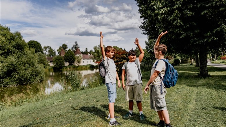 Drei Jungen mit Rucks&auml;cken stehen auf einer Wiese neben einem Teich und heben die Arme in die Luft.