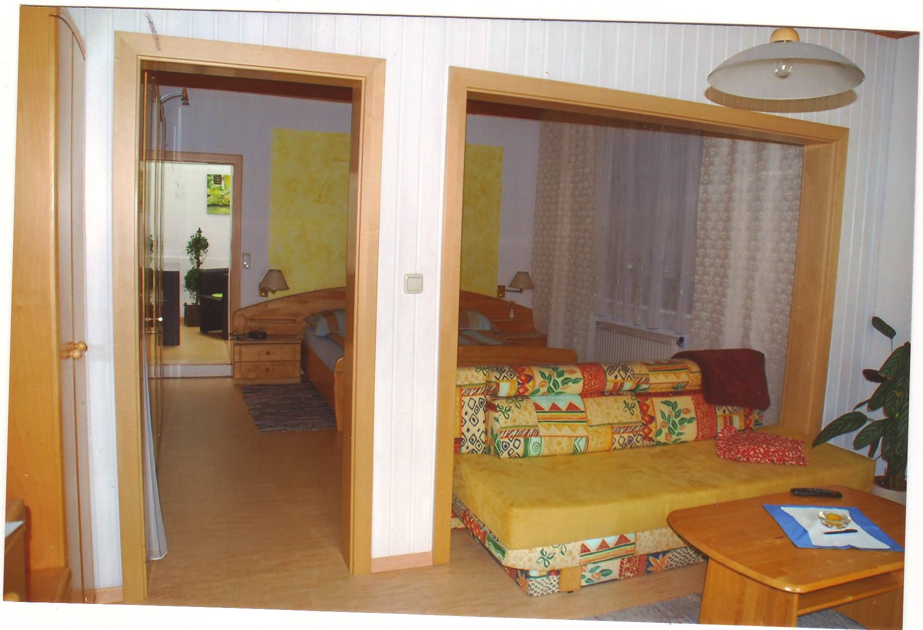 Ein gemütliches Gästezimmer mit buntem Sofa, Holztisch und Blick auf ein Schlafzimmer mit Doppelbett.