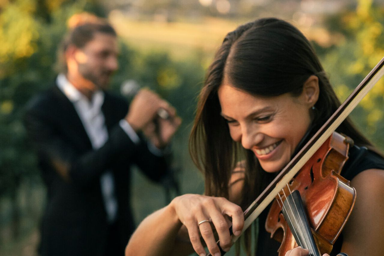 Eine Frau spielt draußen fröhlich Geige, während ein Mann im Hintergrund eins Mikrofon singt. Beide sind elegant gekleidet, und die Umgebung scheint ein weinbergähnliches Setting zu sein.