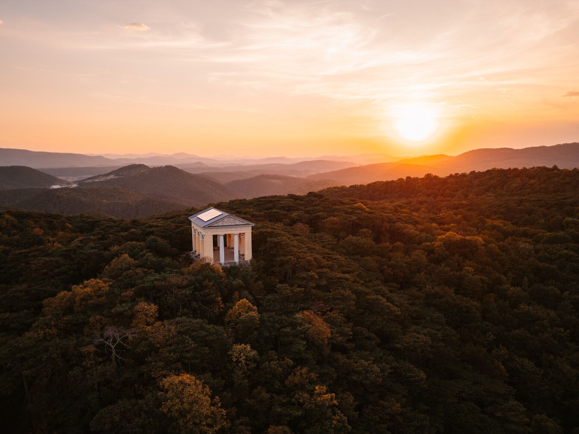 Der Husarentempel thront majestätisch über dem dichten Wald, während die untergehende Sonne den Himmel in ein warmes Gold taucht. Diese idyllische Kulisse lädt dazu ein, die Ruhe der Natur zu genießen und die atemberaubende Aussicht auf die umliegenden Berge zu bewundern.