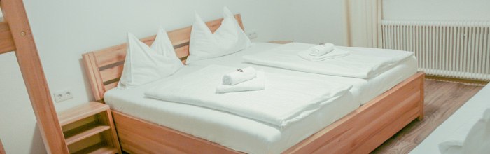 Ein gemütliches Doppelbett in einem hellen Zimmer mit Holzrahmen und weißen Bettlaken, dekoriert mit gefalteten Handtüchern.