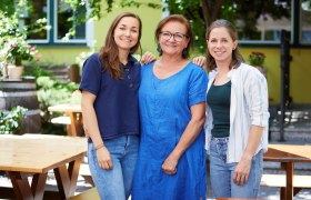 Drei Frauen stehen l&auml;chelnd in einem Garten mit Holztischen und Pflanzen.