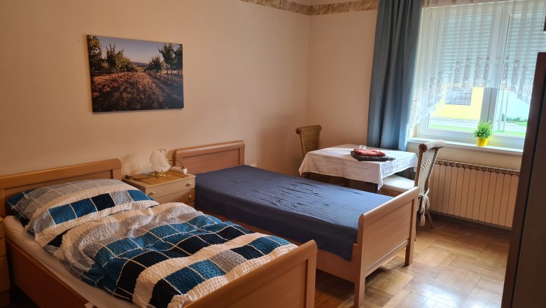 Ein einfach eingerichtetes Schlafzimmer mit zwei Einzelbetten, einem Tisch mit Stuhl und einem Fenster mit Vorh&auml;ngen.